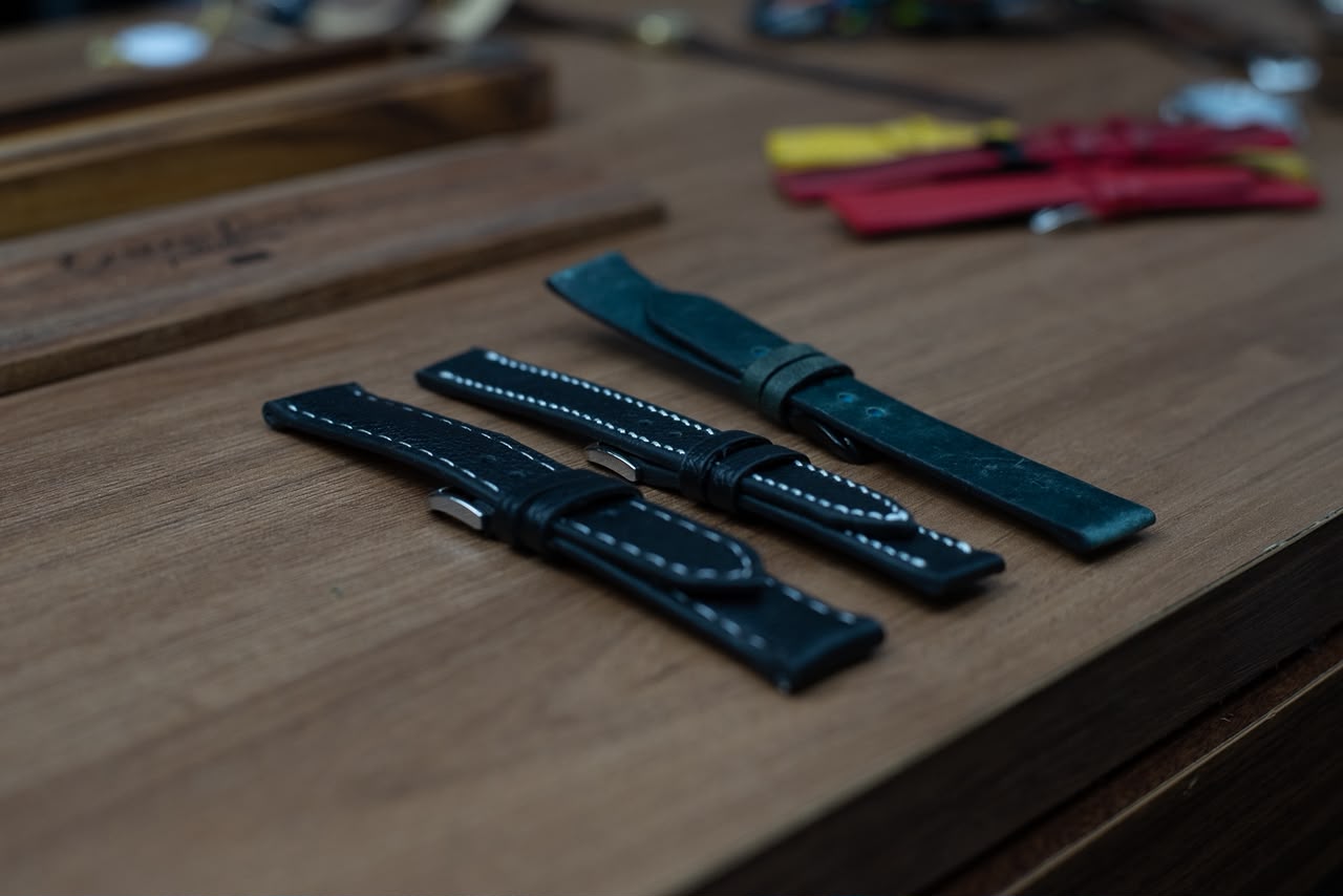 Três pulseiras em tons escuros, preto, preto com costura branca e verde petróleo, sobre a bancada de trabalho
